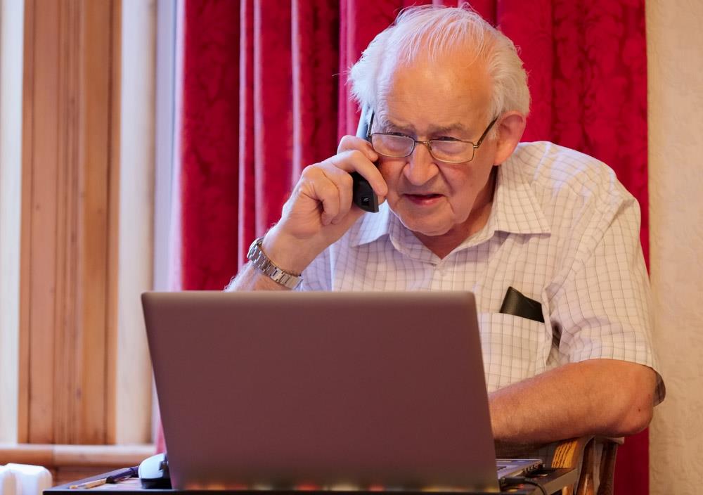old elderly senior man on phone at-laptop computer at risk to cyber attack and online bank fraud